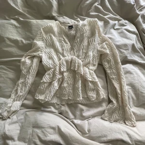 Vit omlottblus med volanger - Säljer en vit omlottblus från Gina med volangdetaljer och långärmad design. Blusen har en v-ringad hals och är gjord i ett lätt, krinklat material som ger en cool textur. Perfekt för dig som gillar romantiska och trendiga plagg.