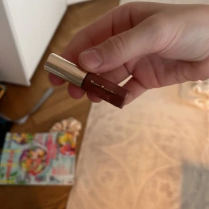 Rare Beauty läppglans i brunrosa nyans - Säljer ett läppglans från Rare Beauty i en snygg brunrosa nyans. Kommer i en liten smidig tub med guldig kork och applikator. Perfekt för att få glansiga och återfuktade läppar med en hint av färg. Liten och enkel att ta med i väskan.