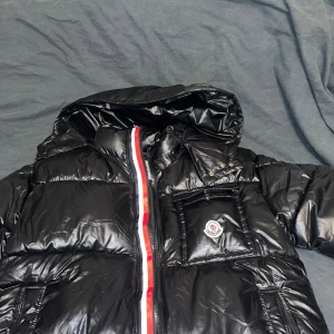 Svart pufferjacka från Moncler - Säljer en svart pufferjacka från Moncler med glansig finish och huva. Jackan har en röd, vit och blå rand längs dragkedjan, en bröstficka med Moncler-logga och flera praktiska fickor. Perfekt för kalla dagar och riktigt snygg streetstil. Använd 1 gång går o skanna på loggan 
