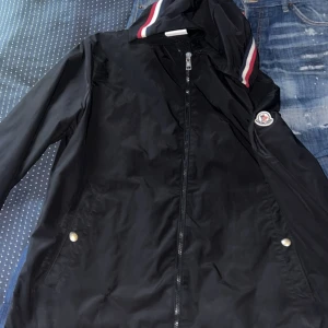 Svart vindjacka från Moncler - Svart vindjacka från Moncler med huva och dragkedja framtill. Jackan har Moncler-logga på ärmen och snygga rödvita detaljer på huvan. Tillverkad i polyester och har praktiska fickor med tryckknappar. Perfekt för dig som vill ha en stilren och sportig look.