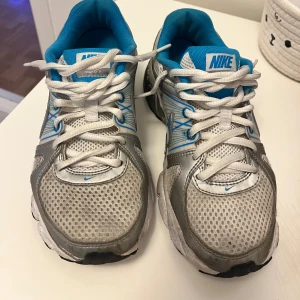 Nike sneakers i silver och blå mesh - PASSAR NÄSTAN ALLT. pris ej hugget i sten.   Nike sneakers med vit och silverfärgad mesh, blå detaljer och vit snörning. Skorna har en sportig vibe med blå Nike-logga på sidan och vadderad blå insida. Sulan är svart och vit med bra grepp, perfekt för dig som gillar en aktiv stil.   