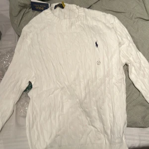 Vit kabelstickad tröja Polo Ralph Lauren - Vit kabelstickad tröja från Polo Ralph Lauren med klassisk rund halsringning och broderad marinblå logga på bröstet. Långärmad modell i mjuk bomull, perfekt för lager-på-lager under höst och vinter.