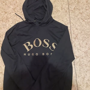 Svart hoodie från Hugo Boss - Svart hoodie från Hugo Boss med stort BOSS-tryck i beige på bröstet. Klassisk huva med dragsko och långa ärmar. Perfekt för dig som vill ha en stilren och bekväm tröja med premiumkänsla.