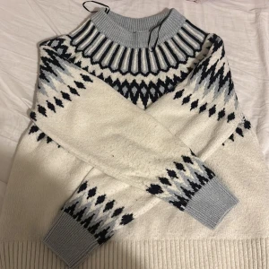 Vit, svart och blå stickad tröja med mönster - Säljer en mysig stickad tröja i vitt med grått och svart mönster över bröst och ärmar. Tröjan har ribbade muddar och hög krage i ljusgrått. Perfekt för kalla dagar och har en klassisk nordisk vibe. Den värmer ordentligt och är som sagt, väldigt mysig.