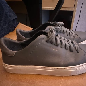 Grå sneakers från Axel Arigato - Snygga grå sneakers från Axel Arigato med minimalistisk design. Skorna har ovandel i slätt skinn, detaljer i mocka vid hälen och vita platta sulor. Klassisk snörning och diskret logga i guld på sidan. Perfekta för en clean och stilren look.