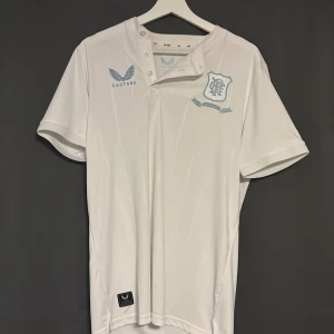 Vit Rangers FC jubileumströja Castore - Säljer en vit fotbollströja från Castore, officiell Rangers FC 150-årsjubileum. Tröjan har korta ärmar, ljusblå detaljer och klubbmärke på bröstet. Materialet är lätt och andas, perfekt för match eller träning. Unik knappdesign vid halsen och diskreta stjärnor på ryggen.