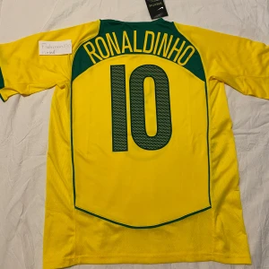 Brasilien Ronaldinho #10 Nike fotbollströja - Gul Brasilien fotbollströja från Nike med gröna detaljer, tryckt nummer 10 och Ronaldinho på ryggen. Klassisk design med landslagets emblem på bröstet och Nike-logga. Aldrig använd.