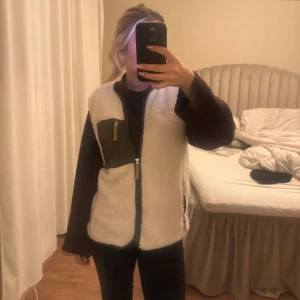 Vit fleeceväst med dragkedja - Säljer en vit fleeceväst med svart kant och en grön bröstficka med dragkedja. Västen har hel dragkedja framtill och är i en loose passform. Perfekt att slänga över en hoodie eller tröja för extra värme. Snygg och enkel design som funkar till många stilar.