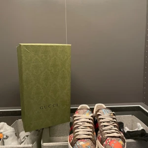 Gucci sneakers med tecknat limiterat design - Säljer ett par unika Gucci sneakers med färgglatt tecknat mönster, klassiska gröna och röda ränder på sidan och orange detalj vid hälen. Skorna har snörning, platt sula och är tillverkade i canvas med skinninslag. Perfekta för dig som vill sticka ut med din stil.