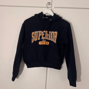 Mörkblå hoodie från H&M med tryck - Mörkblå hoodie från H&M med orange och vitt tryck på bröstet där det står 'SUPERIOR since FOREVER'. Tröjan har luva med snörning, ribbade muddar och är lite kortare än en vanlig hoodie. 