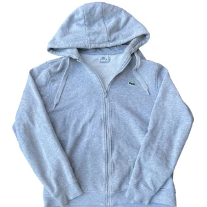 Grå Lacoste Hoodie - Snygg ljusgrå hoodie från Lacoste med klassisk krokodillogga på bröstet. Tröjan har huva med snörning, hel dragkedja och ribbade muddar. Perfekt för en chill och sportig stil. Tillverkad i mjuk bomullsmix för extra komfort. Har hål fram på armarna men inte så märkbara.