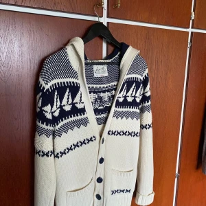 Vit och marinblå stickad kofta med segelbåtsmönster - Säljer en stickad kofta i vitt och marinblått med segelbåtsmönster och knappar framtill. Koftan har huva, långa ärmar och två fickor. Perfekt för kyliga sommarkvällar och har en mysig känsla.  Sällsynt kofta. Finns fåtal av dessa på plick varav jag vet. Pris kan diskuteras vid snabb affär!