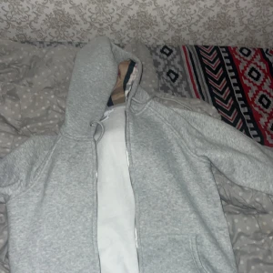 Grå hoodie med dragkedja - Säljer en stilren ljusgrå hoodie med dragkedja och huva. Tröjan har långa ärmar, fickor framtill och ribbade muddar. Perfekt för chill dagar eller när du vill ha en enkel och clean look.