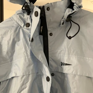 Ljusblå vindjacka från Weather Report - Snygg ljusblå vind/lättare regnjacka från Weather Report med huva och svarta detaljer. Jackan har dragkedja, tryckknappar och justerbar snörning vid halsen. Perfekt för blåsiga dagar och har en clean look med logga på bröstet.