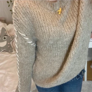 Beige stickad tröja med snörning - Mysig beige stickad tröja med grov struktur och vit snörningsdetalj längs ena ärmen. Tröjan har lång ärm och en avslappnad passform, perfekt för kyliga dagar.