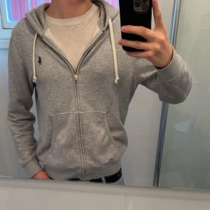 Grå hoodie från Polo Ralph Lauren - Hoodie ifrån Ralph Lauren, Tröjan är i princip helt oanvänd. Skulle säga att tröjan sitter som bäst vid 170-183cm.                               Bara att höra av sig vid funderingar. 