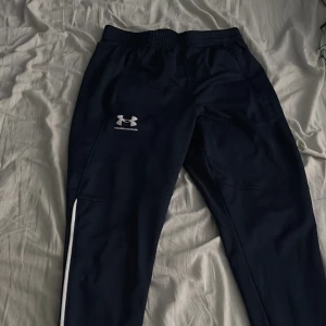 Svarta träningsbyxor från Under Armour - Skickar gärna fler bilder Snygga svarta träningsbyxor från Under Armour med vitt logotryck på låret och vit detalj längs benet. Byxorna har elastisk midja och är perfekta för sport eller chill. Materialet är mjukt och följsamt för maximal rörelsefrihet.