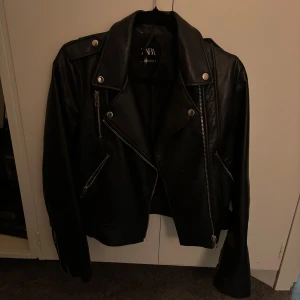 Svart skinnjacka från Zara - Snygg svart skinnjacka från Zara med klassisk bikerstil. Jackan har silvriga dragkedjor, knappar och axelklaffar. Flera fickor framtill och coola detaljer som ger en edgy vibe. Perfekt för dig som vill ha en tidlös och trendig look.
