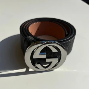 Svart Gucci bälte med GG-spänne - Snyggt svart bälte från Gucci med klassiskt GG-mönster och stort, rundat silverspänne med GG-logga. Bältet är gjort i skinn och har en stilren design som lyfter vilken outfit som helst. Insidan är ljusbrun och bältet har flera hål för justering.