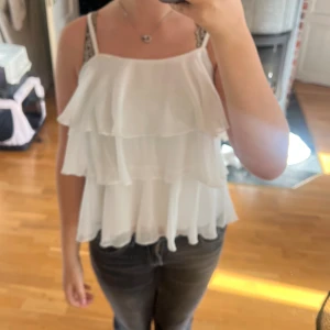 Vit volangig linnetopp med smala axelband - Säljer en superfin vit linnetopp med flera lager volanger och smala axelband. Toppen är luftig och har en lätt, flowy känsla som ger en cool och feminin vibe. Perfekt för varma dagar och att styla med jeans eller kjol.