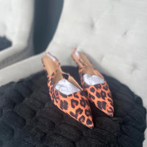 Leopardmönstrade pumps med spetsig tå - Säljer ett par riktigt snygga pumps med leopardmönster i orange och svart. Skorna har spetsig tå, öppen häl och låg klack. Materialet är syntet med slät finish och platt sula undertill. Perfekt för dig som gillar att sticka ut med djurmönster och cool design.