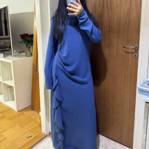 Blå långklänning med volangdetalj - Säljer en blå långklänning med lång ärm och snygg volangdetalj längs sidan. Klänningen har en rak passform och är tillverkad i ett lätt, strukturerat tyg som ger en cool look. Perfekt för dig som gillar stilrena plagg med en twist.