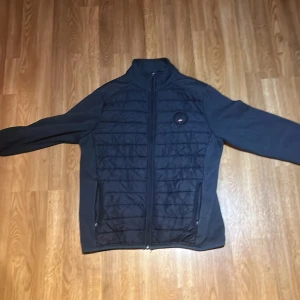 Navy blue cardigan utan hodie - Svart cardigan från Stormy Life med quiltad front och hel dragkedja. Jackan har hög krage, två sidofickor med dragkedja och en liten logga på bröstet. Perfekt för kyliga dagar när du vill ha något lätt men ändå varmt.