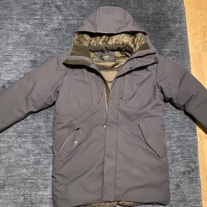 Mörkblå parka jacka från Everest - Säljer en mörkblå parkajacka från Everest med huva och dragkedja. Jackan har två stora fickor framtill, quiltat foder och en clean look. Perfekt för kalla dagar och riktigt skön att bära. Passar dig som gillar stilren och funktionell design.