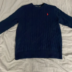 Marinblå kabelstickad tröja Polo Ralph Lauren - Kabelstickad marinblå tröja från Polo Ralph Lauren med klassisk rund hals och lång ärm. Tröjan har det ikoniska röda broderade logot på bröstet och ribbade muddar vid ärmslut och nederkant. Perfekt för en stilren och tidlös look.