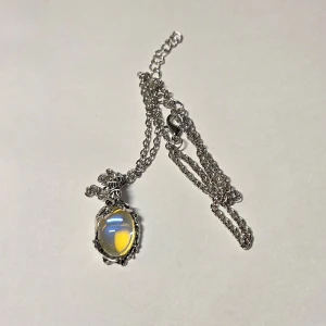 Halsband Färgskiftande StenOpaliserande Månsten/Mood Stone 48 cm Nytt - Magiskt halsband i silverfärg.  Hänget har en unik oval sten som skiftar färg beroende på ljuset och vinkeln, från vitt/blått till gult/orange (liknar en Månsten eller Opalit). Stenens detaljerade infattning i vintage/antik stil ger smycket en mystisk och bohemisk känsla. Perfekt för den som älskar unika  accessoarer och stenar med lyster.  Sten: Skiftande färg / Opaliserande (Mood Stone/Opalit-look) Material: Zink Legering Kedjans längd: Ca 48 cm Stil: Mystisk, Vintage, Bohemian