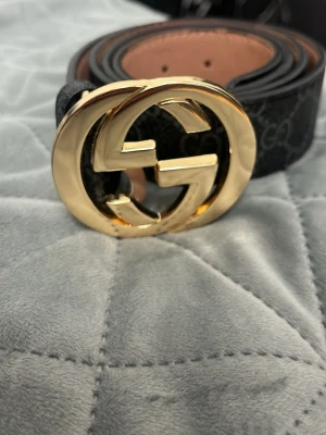  GG Bälte - Säljer ett svart  skärp i skinn med klassiskt GG-mönster och stor guldfärgad GG-logga som spänne. Insidan är beige och har präglad märkning. Perfekt accessoar för att lyfta vilken outfit som helst.