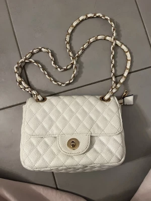 Vit quiltad axelväska med kedja - Säljer en snygg vit axelväska i quiltad design med guldfärgade detaljer. Väskan har en lång guldig kedja med invävt vitt material och en liten ficka på baksidan. Kedjan går att justera. Liten liten defekt på spännet men syns typ inte! 