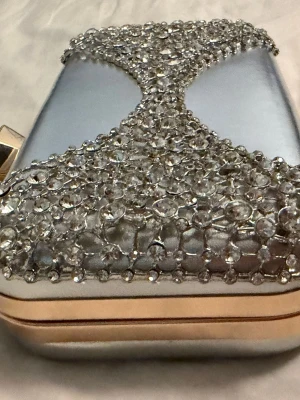 Silver kuvertväska med strassdetaljer - Snygg kuvertväska i metallic silver med glittrande strass framtill i ett coolt mönster. Väskan har guldfärgade kanter och ett stort gnistrande spänne. Perfekt för dig som vill sticka ut och addera extra bling till din look.