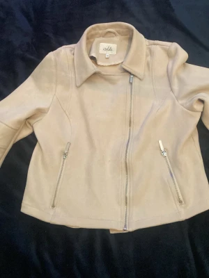 Beige mockajacka från Isolde - Snygg beige mockajacka från Isolde med asymmetrisk dragkedja och två snedställda fickor med dragkedjor. Jackan har markerad krage och coola sömdetaljer på ärmarna. Perfekt för dig som vill ha en stilren och trendig look.