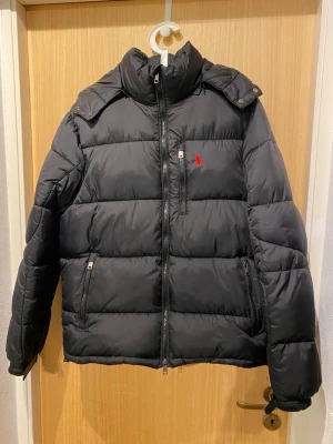 Svart pufferjacka från Polo Ralph Lauren - Säljer en svart pufferjacka från Polo Ralph Lauren i storlek M  med huva och dragkedja framtill. Jackan har en röd logga på bröstet, flera fickor med dragkedja och en hög krage. Perfekt för kalla dagar. är osäker om den är äkta👍😁