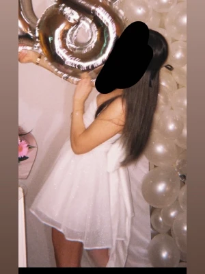 Vit glittrig axelbandslös kortklänning - Supersöt vit axelbandslös klänning med glittrigt tyg och stor rosett i ryggen. Kjolen är fluffig och har flera lager för extra volym. Perfekt för dig som vill sticka ut på bal eller student. Materialet känns lätt och skimrande.