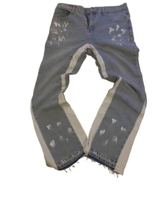 Blå jeans med beige paneler och slitningar - Unika blå jeans med breda beige paneler längs benen och slitna detaljer framtill och baktill. Råa, fransiga benslut ger en cool edge. Jeansen har klassisk femficksdesign och är tillverkade i denim. Perfekta för dig som vill sticka ut med en streetwear vibe.