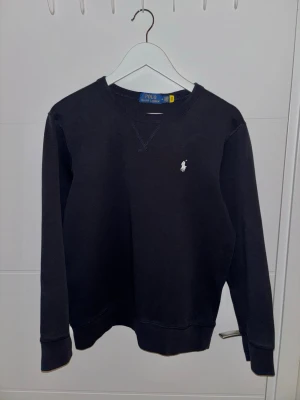 POLO RALPH LAUREN – Mörkblå sweatshirt i storlek M - Säljer en högklassig och eftertraktad mörkblå sweatshirt från Polo Ralph Lauren. Den är i storlek M och har den klassiska, tidlösa polokragen och den ikoniska hästen/loggan. Plagget är tillverkat av tjock, mjuk och hållbar bomull av premiumkvalitet, vilket märks på både känsla och hållbarhet.  Sweatshirten är i mycket gott skick, men eftersom den har blivit för liten för mig får den ett nytt hem. Inga synliga skador eller fläckar.  Bor i Strängnäs. Produkten finns redo för upphämtning. Kan skick