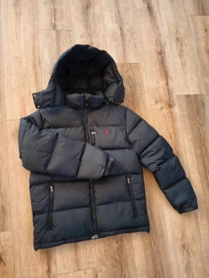 Svart pufferjacka från Polo Ralph Lauren - Säljer en svart pufferjacka från Polo Ralph Lauren med röd logga på bröstet. Jackan har huva, två dragkedjeförsedda fickor och kraftig vaddering som håller dig varm. Klassisk rak passform och hel dragkedja framtill.