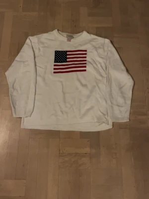 Vit tröja med amerikansk flagga - Vit långärmad tröja med en stor amerikansk flagga broderad på bröstet. Klassisk rund halsringning och mjukt material som känns skönt mot huden. Perfekt för dig som vill ha en enkel men ändå statement tröja.