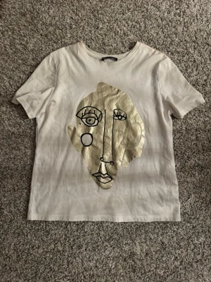Vit t-shirt med guldtryck från Zara - Vit t-shirt från Zara med korta ärmar och ett stort abstrakt ansikte i guld och svart på framsidan. Skön passform och tillverkad i mjuk bomull. Perfekt för dig som gillar unika prints och vill sticka ut lite extra.