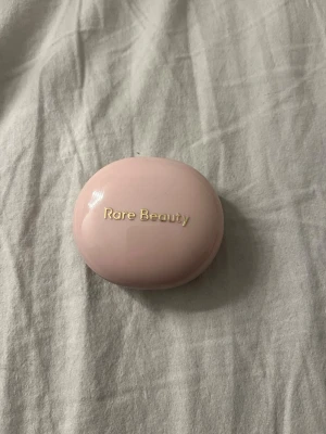 Rare Beauty Nearly Mauve Cream Blush - Rare Beauty Nearly Mauve är ett krämrouge i en söt, ljusrosa rund dosa med spegel. Färgen är en mjuk mauve-ton som ger kinderna en naturlig och fräsch look. Perfekt att ha i väskan för touch-ups under dagen.