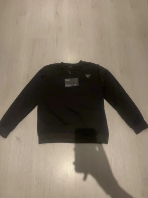 Svart sweatshirt från Prada - Snabb leverans. Säljer en svart sweatshirt från Prada med klassisk rund hals och diskret logotyp-patch på bröstet. Tröjan har långa ärmar och ribbade muddar vid ärmslut och nederkant. Tillverkad i mjuk bomull för en clean och stilren look. Det står L men känns som M