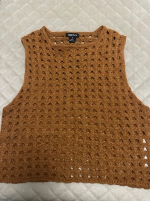 Brun stickad ärmlös topp från Boohoo - Säljer en brun stickad ärmlös topp från Boohoo i storlek M. Toppen har ett hålmönster över hela och är croppad, perfekt för lager-på-lager eller till sommaren. Superenkel att styla med jeans eller kjol för en chill vibe.
