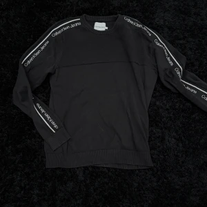 Svart tröja från Calvin Klein Jeans - Svart långärmad tröja från Calvin Klein Jeans med logotyp längs ärmarna. Tröjan har ribbade muddar vid ärmslut och nederkant samt en klassisk passform. Perfekt för en stilren och avslappnad look.