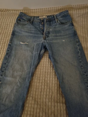 Ljusblå jeans från Jack & Jones - Säljer ett par ljusblå jeans från Jack & Jones med klassisk femficksdesign och knäppning med knappar. Jeansen har en lätt sliten look med en liten reva på låret och är tillverkade i bomull. Perfekta för dig som gillar en avslappnad och tidlös stil.