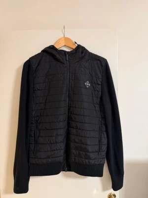 J.Lindeberg Cardigan - Säljer en svart cardigan från J.Lindeberg i storlek M. Tröjan har quiltad framsida och rygg, stickade ärmar och ribbade muddar. Utrustad med dragkedja framtill, fickor och en schysst huva. Perfekt för kyliga dagar när du vill vara både varm och stilren.