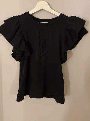 Svart volangblus från Zara - Supersnygg svart blus från Zara med breda volangärmar och ribbad struktur. Toppen har rund halsringning och är gjord i ett mjukt, lätt material som ger en lyxig känsla. Perfekt för dig som vill ha något extra till din outfit.