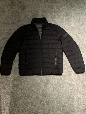 Svart pufferjacka från Marc O'Polo - Säljer en svart pufferjacka från Marc O'Polo med horisontella sömmar och hög krage. Jackan har dragkedja framtill och två sidofickor med dragkedja. Perfekt för kyliga dagar och har en clean, stilren look.
