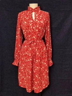 Röd blommig midiklänning med knytning - Supersnygg röd klänning med vitt blommönster, hög krage och nyckelhålsöppning fram. Knyt i midjan för en markerad siluett och långa ärmar med volang vid manchetterna.